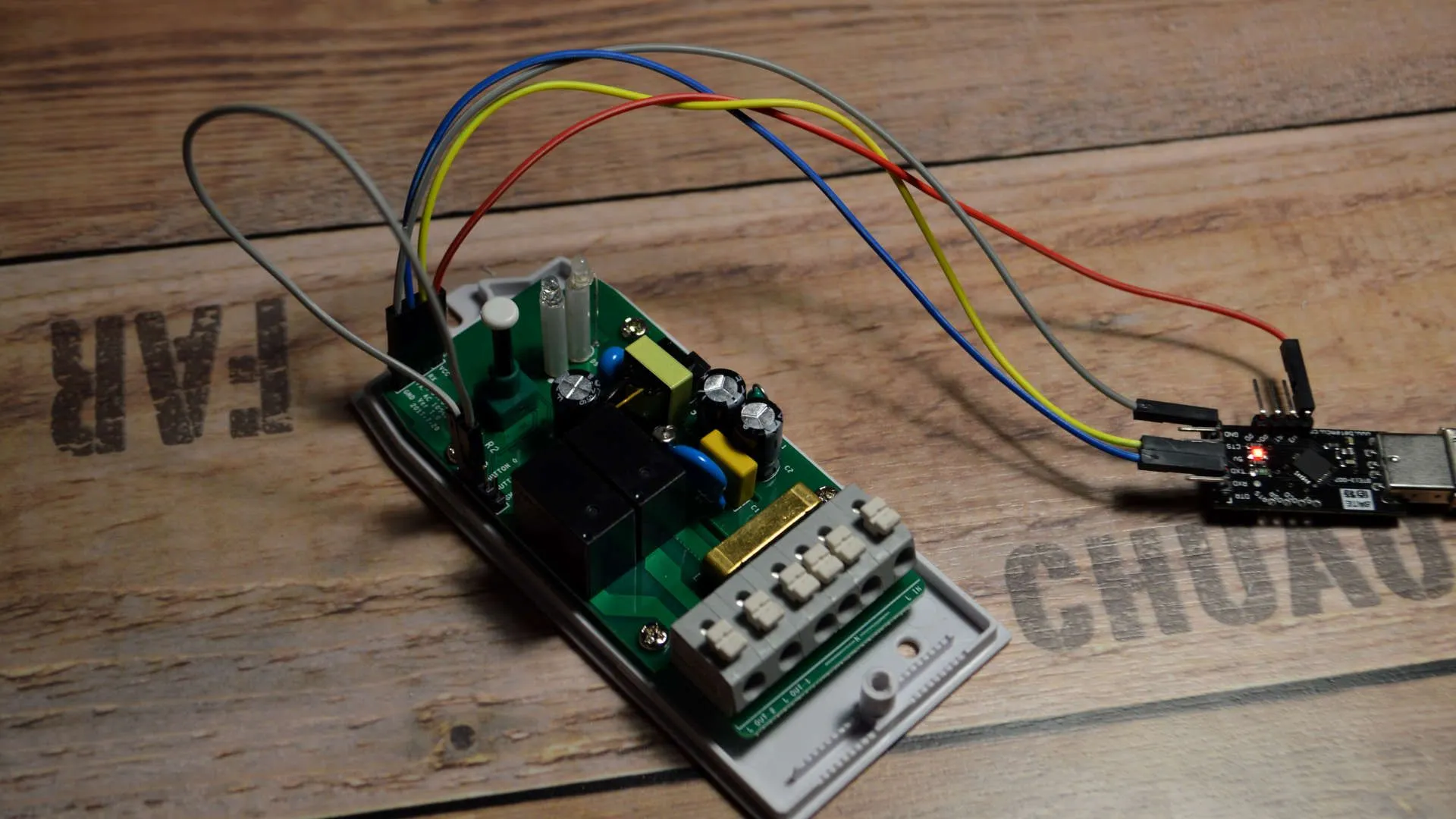 The Sonoff Dual circuit board with its casing open on a wooden surface, connected via gray, blue, red, and yellow jumper wires to a USB-to-serial programmer adapter with a lit red LED, used for flashing custom firmware.