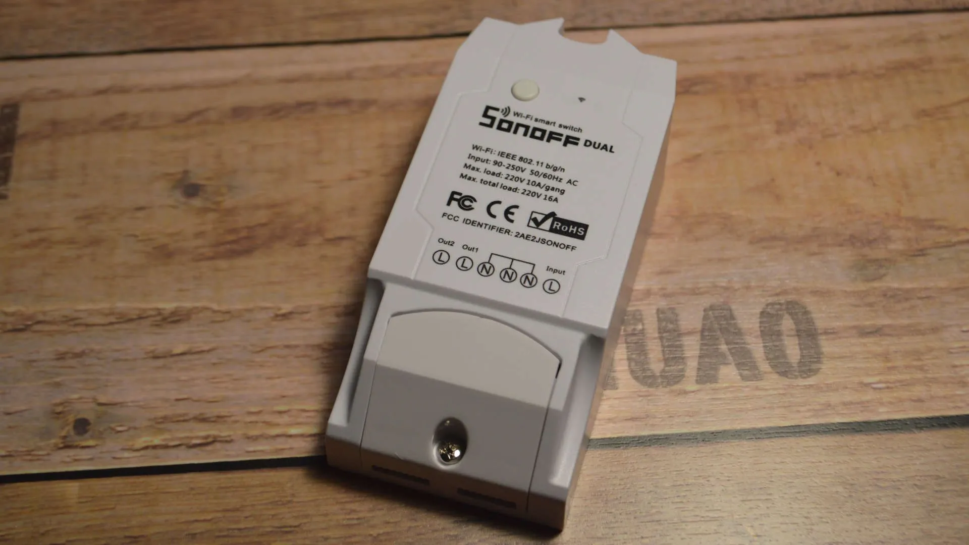 A Sonoff Dual Wi-Fi smart switch lying on a wooden surface, showing its white plastic casing with the Sonoff Dual label, FCC/CE/RoHS certification marks, wiring diagram with Out2 L, Out1 L, N, N, N, and Input L terminals, and a single button on top.