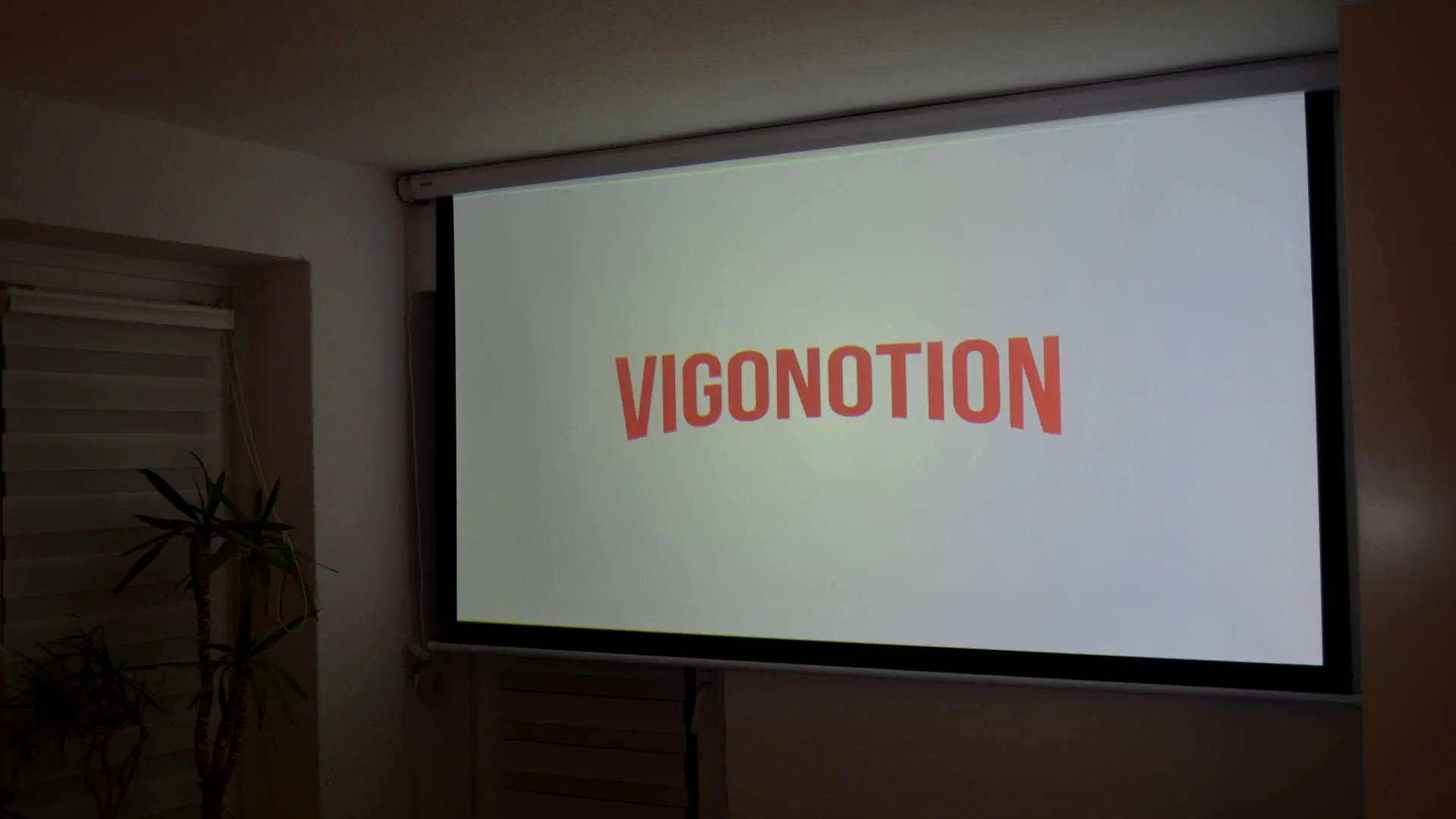 A motorized projection screen lowered in a living room, displaying the word "VIGONOTION" in bold orange letters, with a balcony door visible to the left