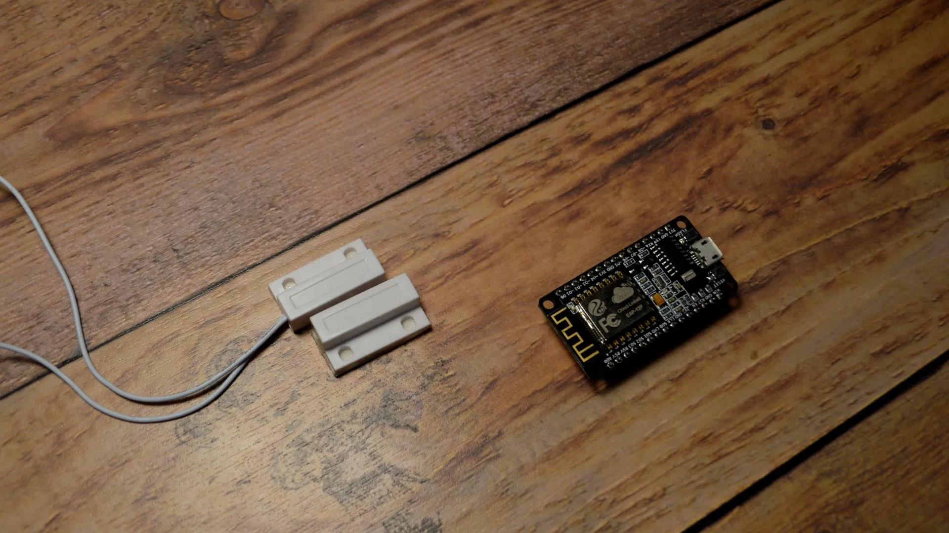 A white two-part magnetic reed contact sensor with a grey wire lying next to a black NodeMCU V1.0 ESP8266 board with gold pin headers, both placed on a wooden surface