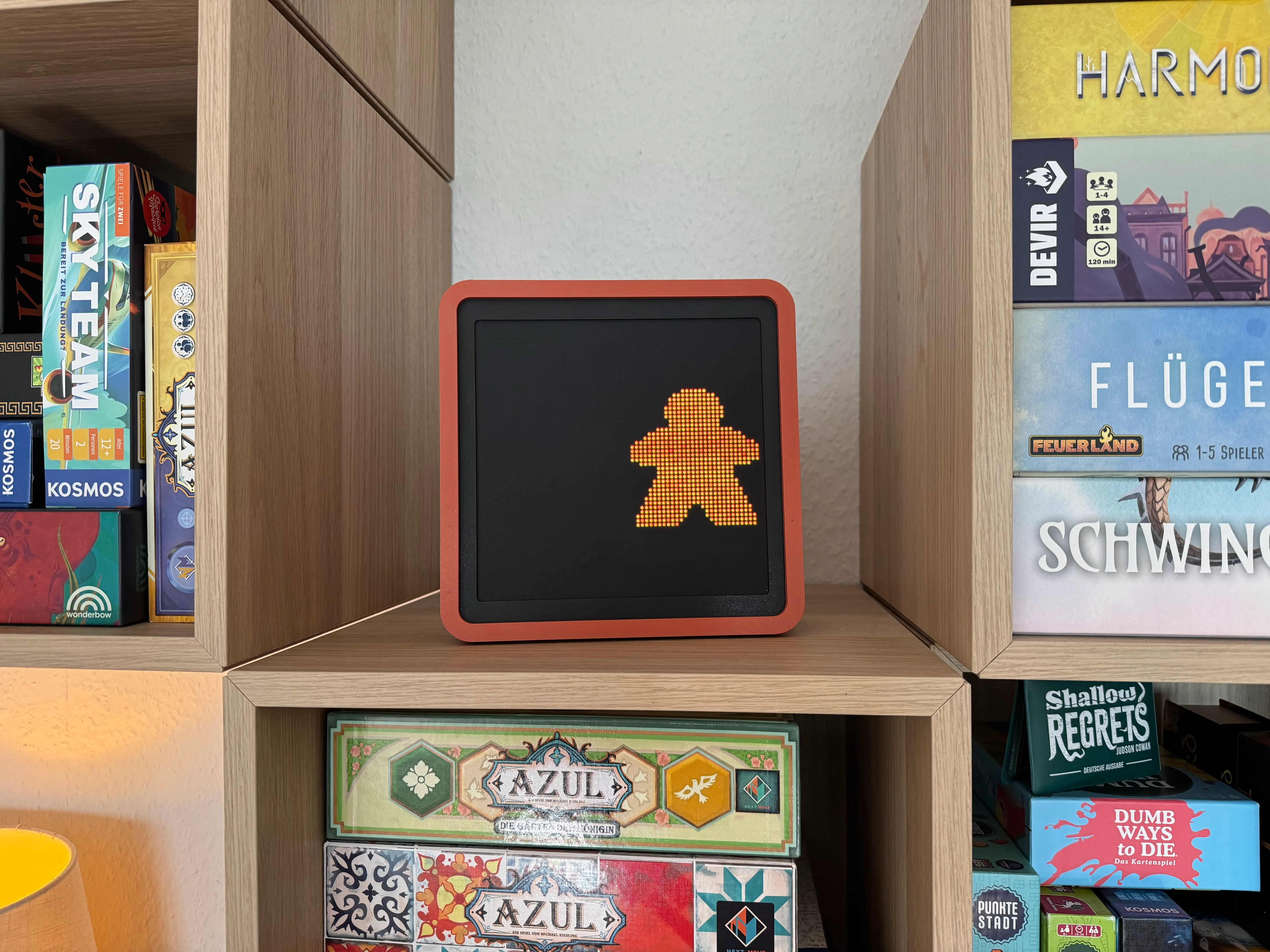 The finished Meeple Traffic Light standing on a wooden shelf between board games, displaying a glowing orange meeple walking figure on its black LED matrix inside an orange-rimmed case.
