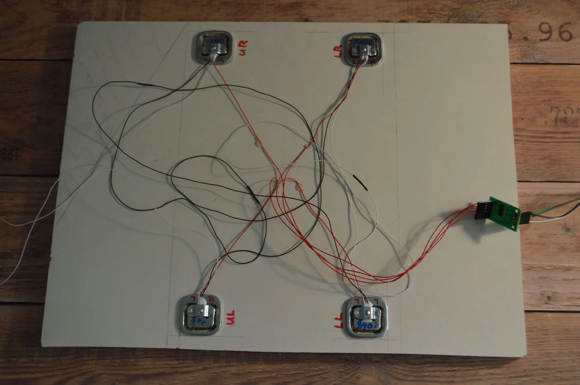A homemade scale platform: a white rectangular board on a wooden floor with four square load cells mounted at each corner, labeled UR, LR, UL, and LL in red marker, connected by a tangle of red, black, and white wires leading to a small green HX711 circuit board on the right edge