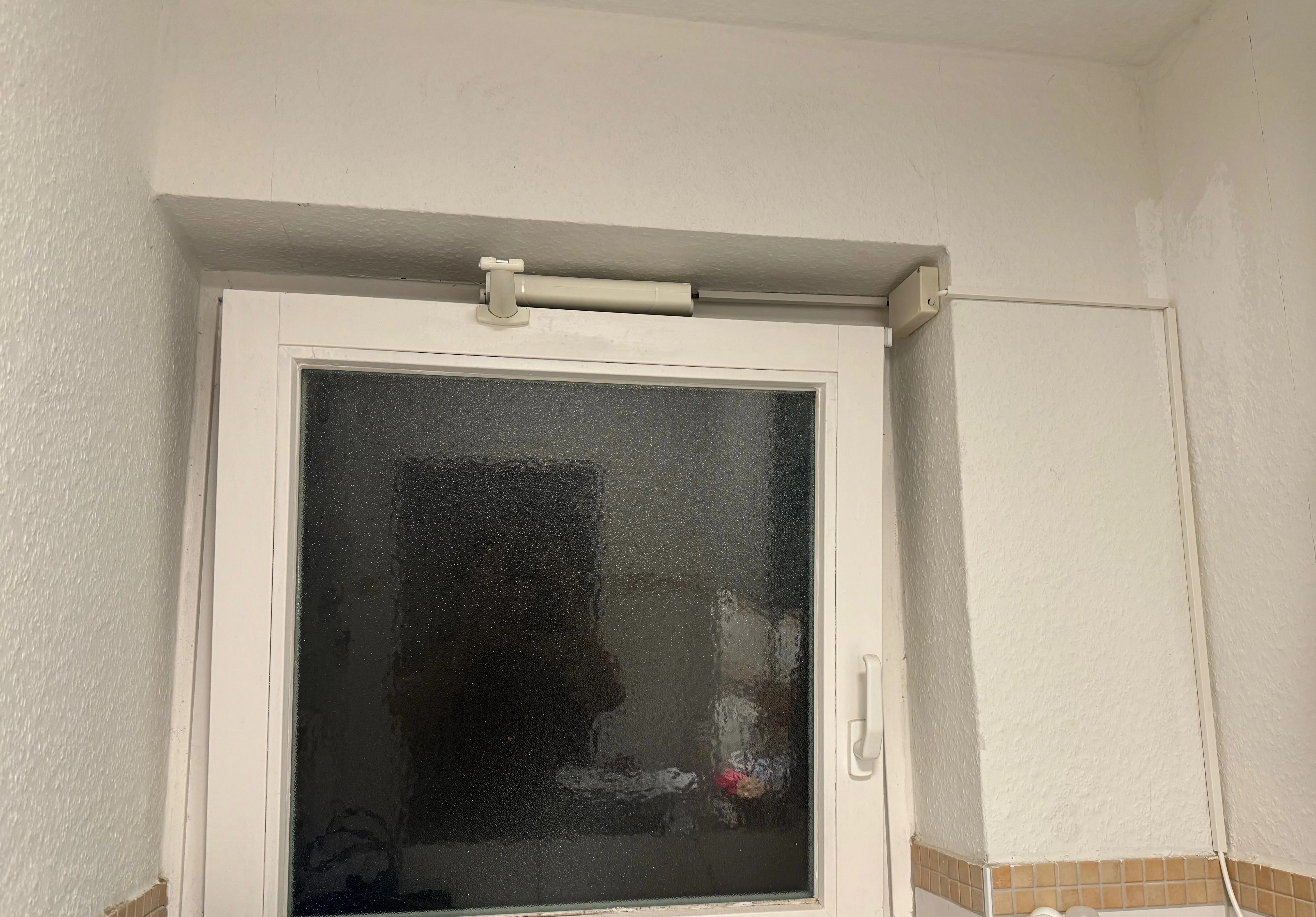 A frosted-glass bathroom window in a white tiled room, with the white cylindrical window opener motor mounted horizontally across the top of the window frame and a white cable running down the right side of the wall